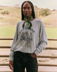 A person with long braids and a green bandana around their neck stands outdoors. They are wearing The Great Inc.'s vintage-style College Sweatshirt, which features an illustration of a cowboy riding a horse and the words "The Great" on it. The background showcases a fenced grassy field and hills, evoking handcrafted in Los Angeles charm.