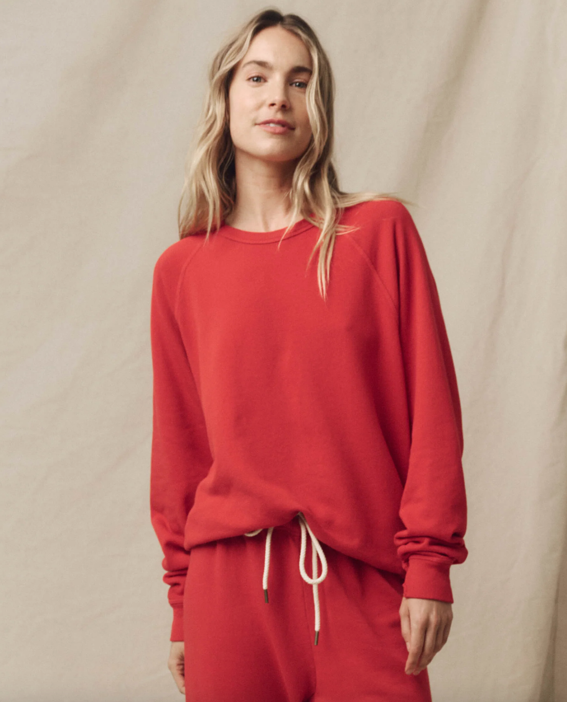 A person with long, wavy hair stands in front of a beige background, dressed in a matching red vintage sweatshirt and pants from The Great Inc. They look at the camera with a neutral expression, hands tucked into the pockets of The College Sweatshirt. A Wild Panther Graphic on the outfit adds an edgy touch.