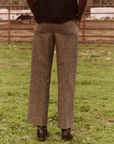 A person is standing on green grass, facing a red metal fence, wearing The Maker Trouser by The Great Inc., which features a gray, lightweight wool blend styled like vintage menswear trousers, and black shoes. The person's hands are in their pockets, with a blurred view of a field and a few trees in the background.