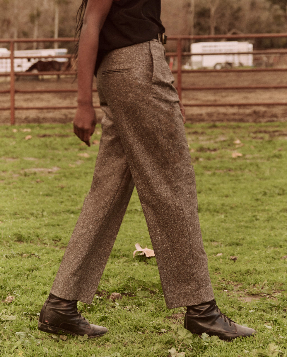 A person walking in a grassy area is seen wearing The Maker Trouser from The Great Inc., paired with a black top and black boots. The blurred background reveals a red metal fence and trees, while the person's head is out of the frame.