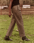 A person walking in a grassy area is seen wearing The Maker Trouser from The Great Inc., paired with a black top and black boots. The blurred background reveals a red metal fence and trees, while the person's head is out of the frame.