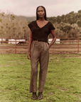 A person stands outside on grass, wearing a black short-sleeve top, gray high-waisted The Maker Trouser by The Great Inc., and black ankle boots. They have one hand in their pocket, and a fence, trees, and hills are visible in the background. The sky is overcast.