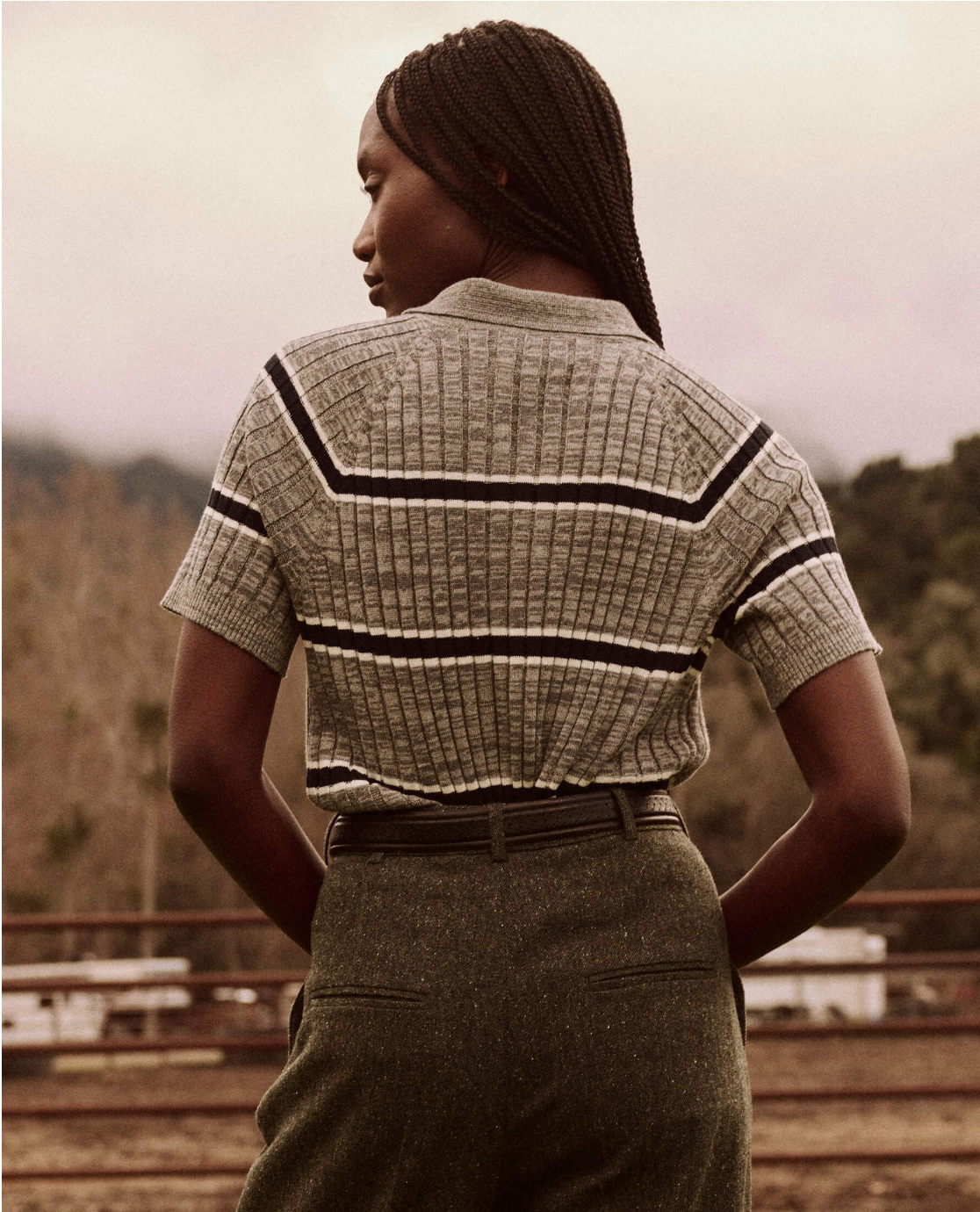 A person with long braided hair stands outdoors, facing away. They are wearing The Classic Polo Sweater by The Great Inc. in heather grey, with fine-gauge rib stripes and high-waisted green pants. The background features wooden fencing and an overcast sky with trees and a mountain in the distance.