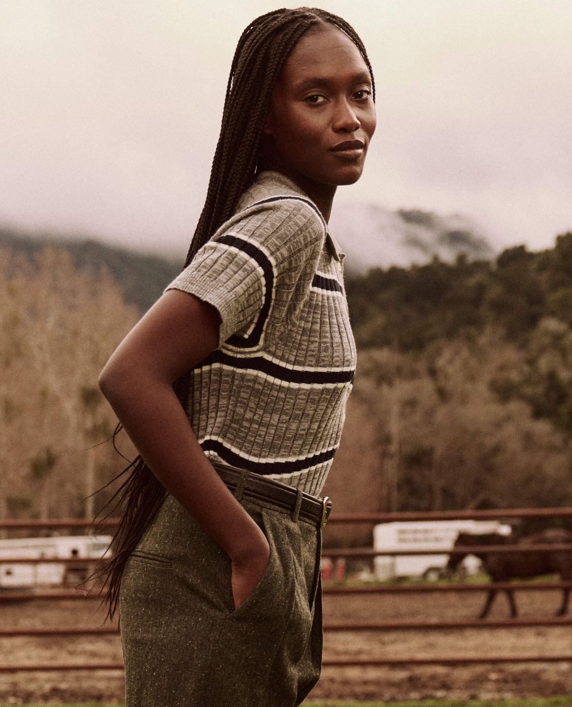 A woman with long braids, wearing The Classic Polo Sweater from The Great Inc., along with high-waisted pants, stands outdoors in a rustic setting featuring mountains, trees, and horse trailers in the background. She has her hands in her pockets and looks confidently at the camera.