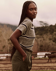 A woman with long braids, wearing The Classic Polo Sweater from The Great Inc., along with high-waisted pants, stands outdoors in a rustic setting featuring mountains, trees, and horse trailers in the background. She has her hands in her pockets and looks confidently at the camera.