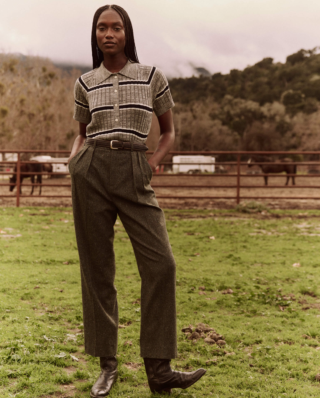 A person with long braided hair stands outdoors on a grassy field, wearing *The Classic Polo Sweater* by *The Great Inc.*, paired with high-waisted trousers. With their hands in their pockets and black boots grounding the vintage prep look, trees and mountains form the idyllic backdrop.