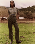 A person with long braided hair stands outdoors on a grassy field, wearing *The Classic Polo Sweater* by *The Great Inc.*, paired with high-waisted trousers. With their hands in their pockets and black boots grounding the vintage prep look, trees and mountains form the idyllic backdrop.