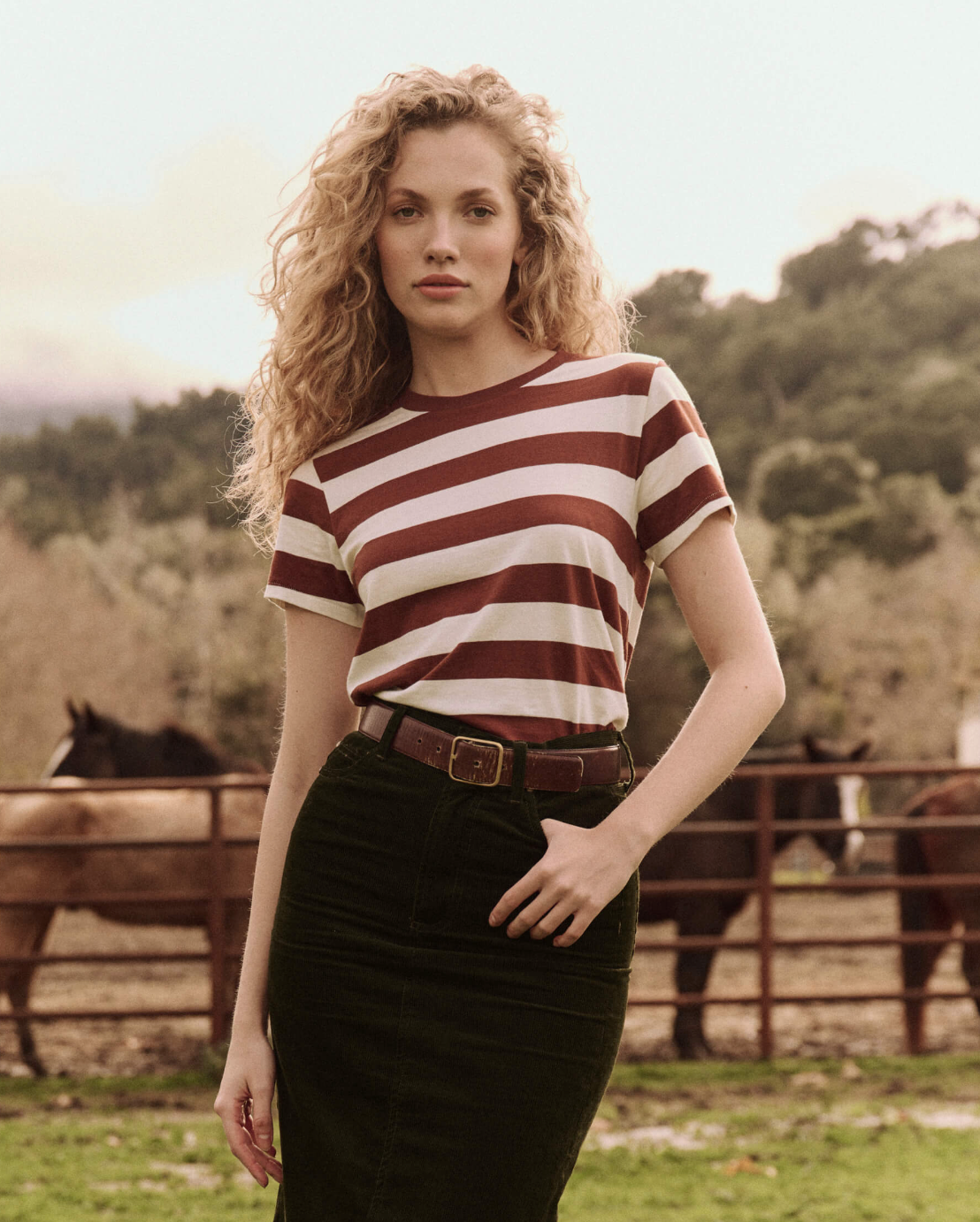 A person with long curly hair is wearing The Great Inc.&#39;s The Little Tee and a dark skirt, standing in a fenced outdoor area with horses in the background. Trees and mountains are visible in the distance.