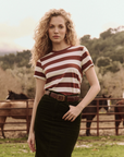 A person with long curly hair is wearing The Great Inc.'s The Little Tee and a dark skirt, standing in a fenced outdoor area with horses in the background. Trees and mountains are visible in the distance.