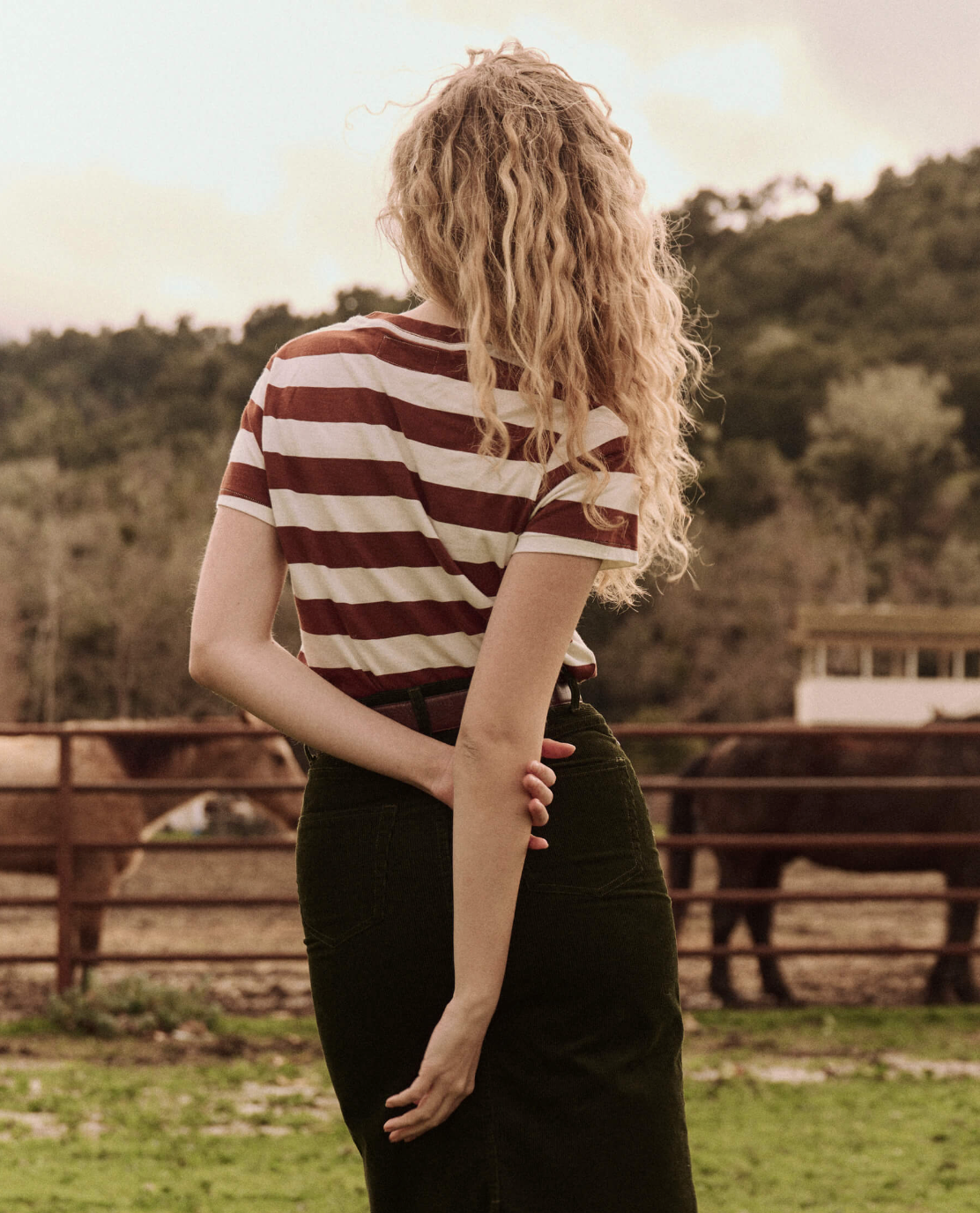 A person with long, curly blonde hair stands with their back to the camera, wearing The Little Tee by The Great Inc. and a skirt. One hand rests on their back. They are in a rural setting with horses and fencing visible in the background.