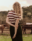 A person with long, curly blonde hair stands with their back to the camera, wearing The Little Tee by The Great Inc. and a skirt. One hand rests on their back. They are in a rural setting with horses and fencing visible in the background.