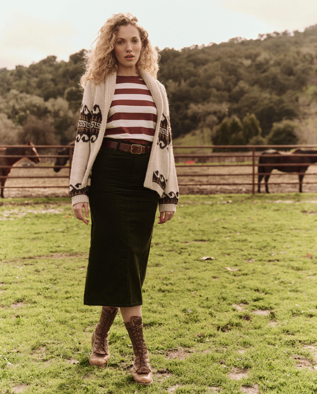 A woman stands in a grassy field with horses in the background. She is wearing a long dark green skirt, The Little Tee by The Great Inc., a beige cardigan with a pattern, and brown lace-up boots. Her hair is curly and blond, and there are green hills behind her.