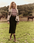 A woman stands in a grassy field with horses in the background. She is wearing a long dark green skirt, The Little Tee by The Great Inc., a beige cardigan with a pattern, and brown lace-up boots. Her hair is curly and blond, and there are green hills behind her.