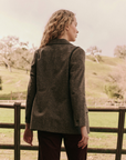 A person with long, wavy hair, wearing The Double Breasted Blazer by The Great Inc., stands with their back to the camera, looking towards a rustic, green hillside. The scene includes a wooden fence and scattered trees in the background under a cloudy sky.