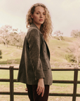 A person with curly blond hair, wearing The Great Inc.'s Double Breasted Blazer in a lightweight wool blend gray fabric and paired with dark pants, stands outdoors near a wooden railing. The green grassy hills and scattered trees form the backdrop as the person looks back over their shoulder with a neutral expression.