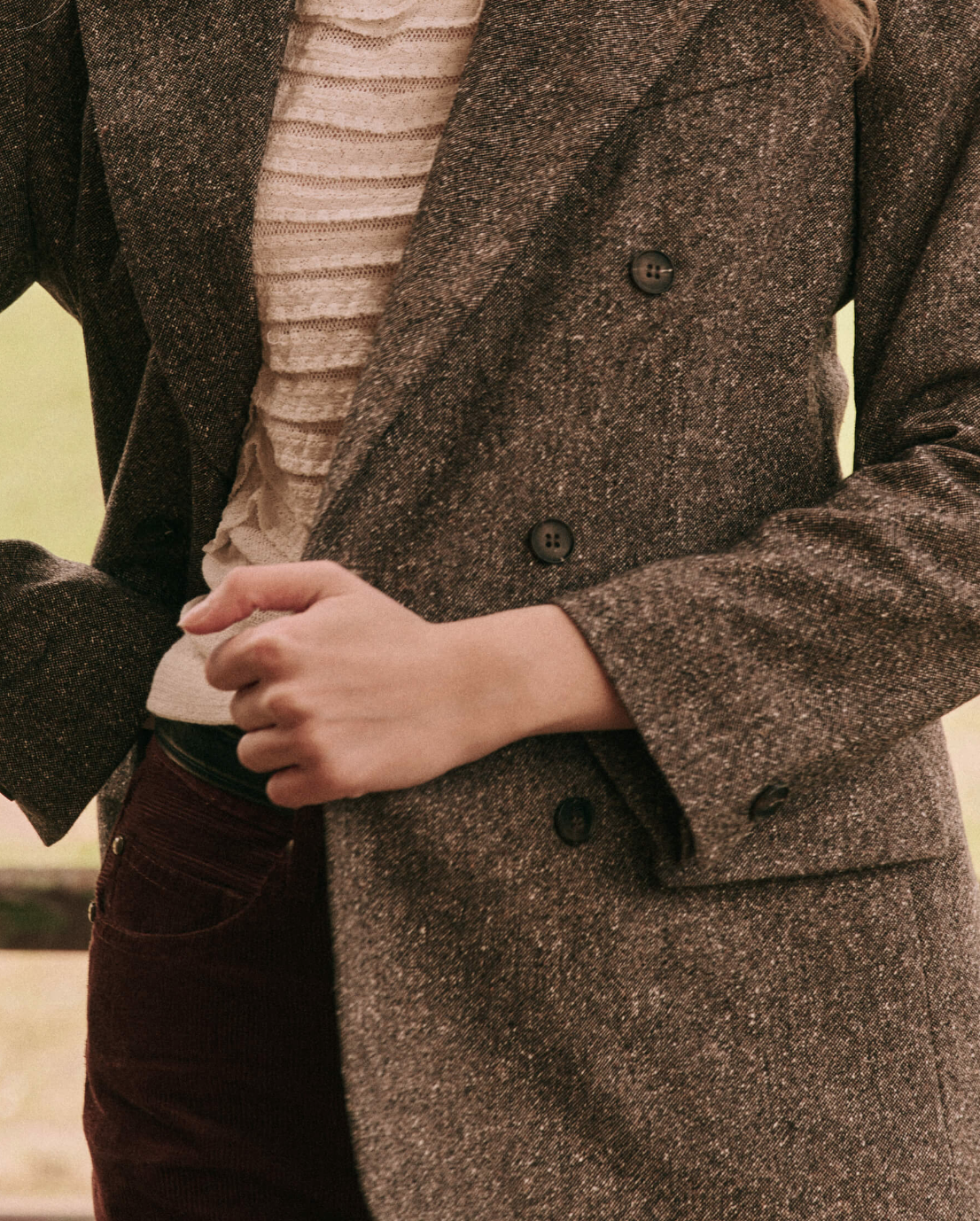 The person is wearing 'The Double Breasted Blazer' by The Great Inc. in gray over a white textured top and dark pants with a slightly visible side pocket. The background features an outdoor setting with a soft, blurred nature view. The person's hands are adjusting the blazer button, showcasing its slim tailored fit.