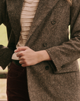 The person is wearing 'The Double Breasted Blazer' by The Great Inc. in gray over a white textured top and dark pants with a slightly visible side pocket. The background features an outdoor setting with a soft, blurred nature view. The person's hands are adjusting the blazer button, showcasing its slim tailored fit.