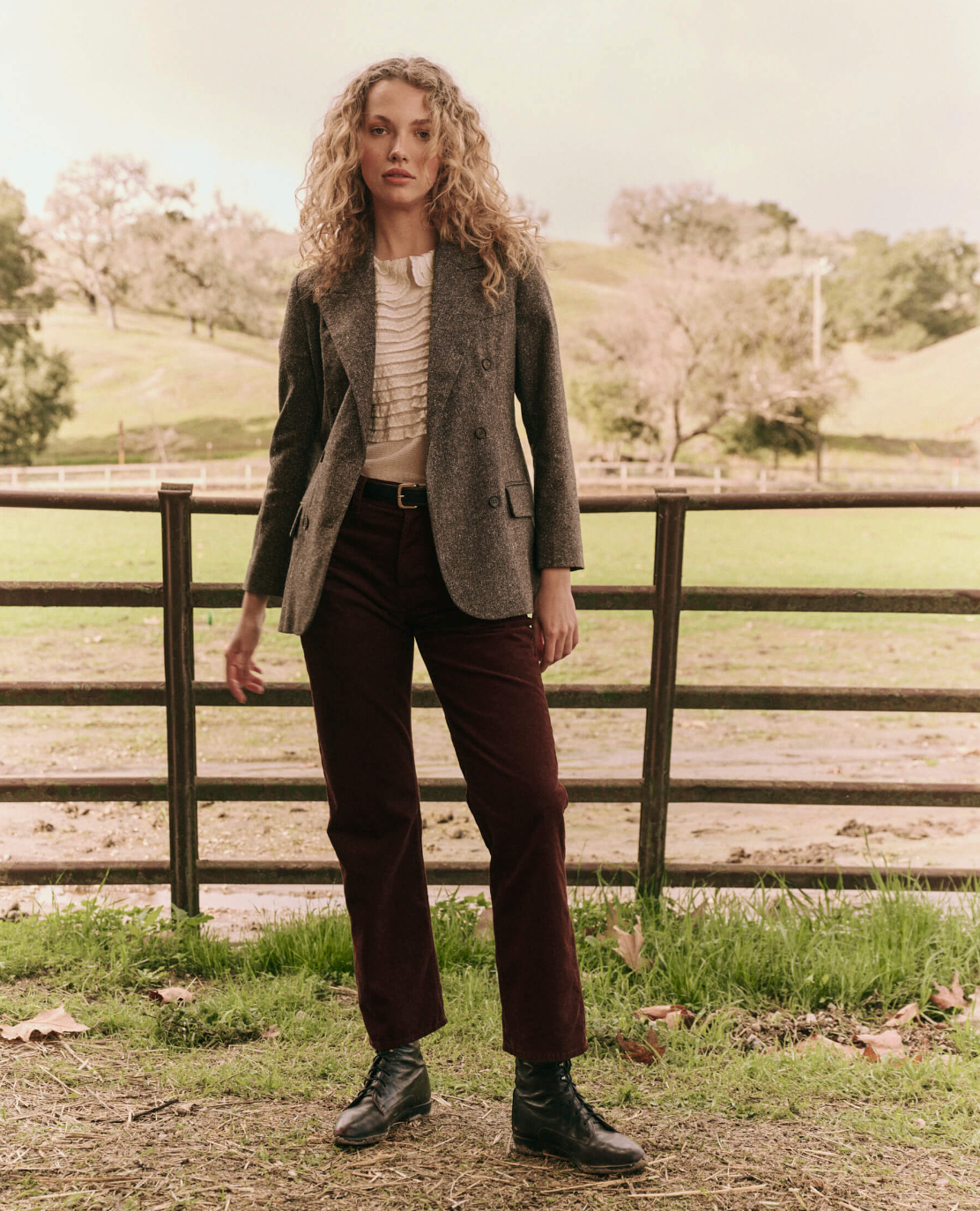 A person with curly hair is standing in front of a wooden fence in an outdoor setting. They are wearing "The Double Breasted Blazer" by The Great Inc. over a white top, burgundy pants, and black boots. The background features green grass, trees, and a slightly cloudy sky.