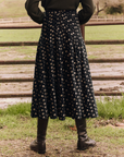 A person stands facing a wooden fence, wearing The Rhythm Skirt by The Great Inc. in black with a small yellow floral pattern reminiscent of the Night Lily Print, a dark green top, and brown leather boots. The background features a grassy field and trees.