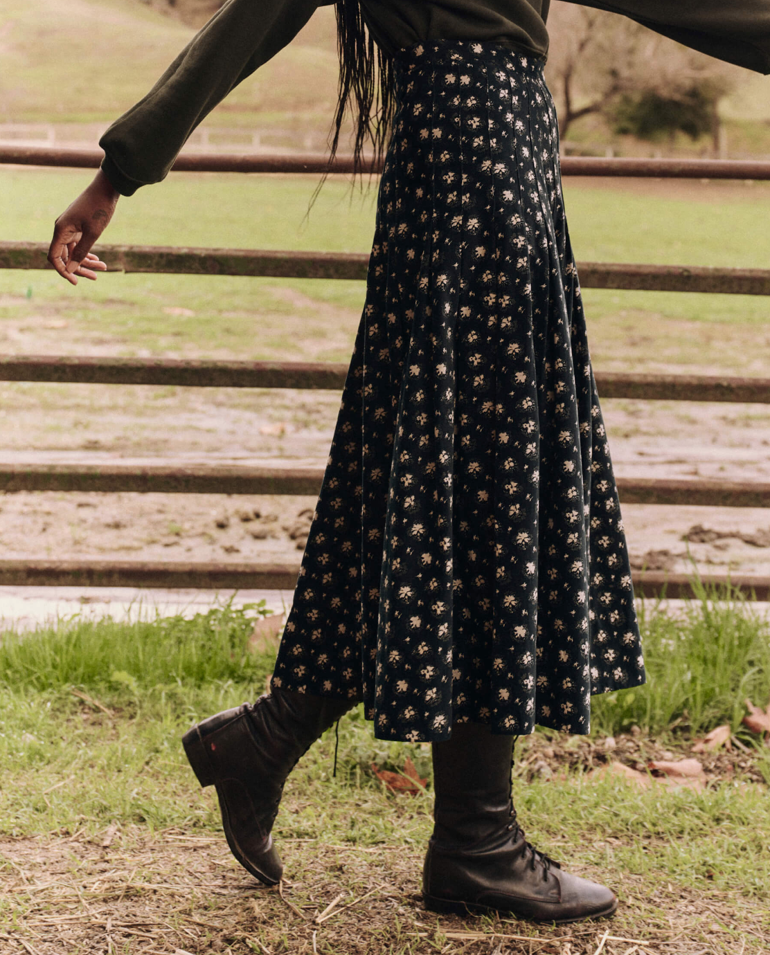 A person wearing a dark green sweater, The Rhythm Skirt—a long black floral midi skirt by The Great Inc.—and black boots is standing on a dirt path with green grass in the background, holding a wooden fence with one hand. The scene suggests a rural or countryside environment.