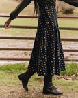 A person wearing a dark green sweater, The Rhythm Skirt—a long black floral midi skirt by The Great Inc.—and black boots is standing on a dirt path with green grass in the background, holding a wooden fence with one hand. The scene suggests a rural or countryside environment.