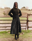 A person stands outdoors in front of a metal gate and grassy field. They wear The Rhythm Skirt from The Great Inc., a dark green sweater, and black boots. Their hair is styled in long braids, and they strike a confident pose with their hands on their hips.