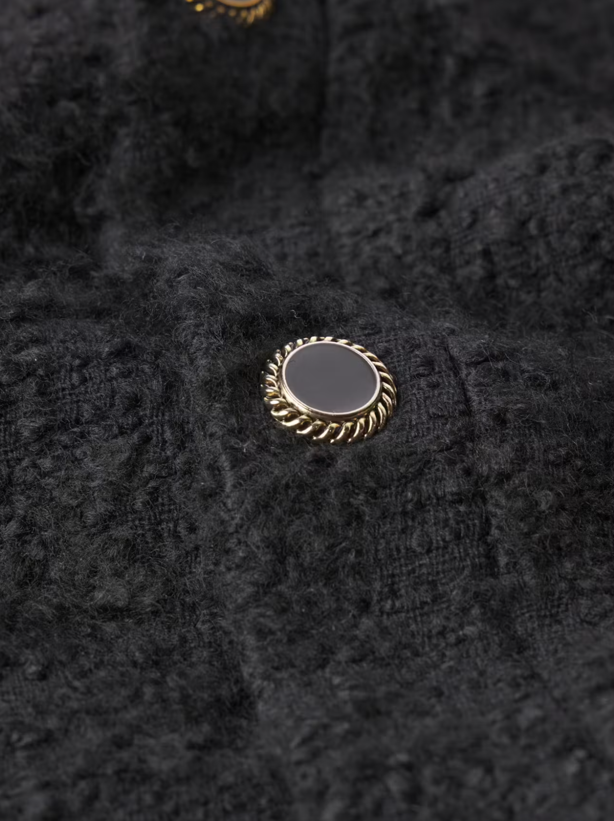 Close-up of the textured black fabric from the Cocoon Short Coat by FRAME, made from a cotton-wool blend. It features a round, black button with an intricate, twisted gold rim that beautifully contrasts with the dark material.