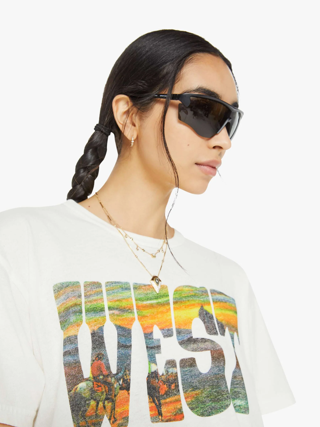 A person with long braided hair and sunglasses, adorned with gold jewelry, shows off an oversized fit graphic white t-shirt called "The Sleep Over" by Mother, made from 100% cotton. The shirt showcases the word "WEST" in colorful letters, reminiscent of a lively Western sunset scene. They strike a pose against a plain white background.