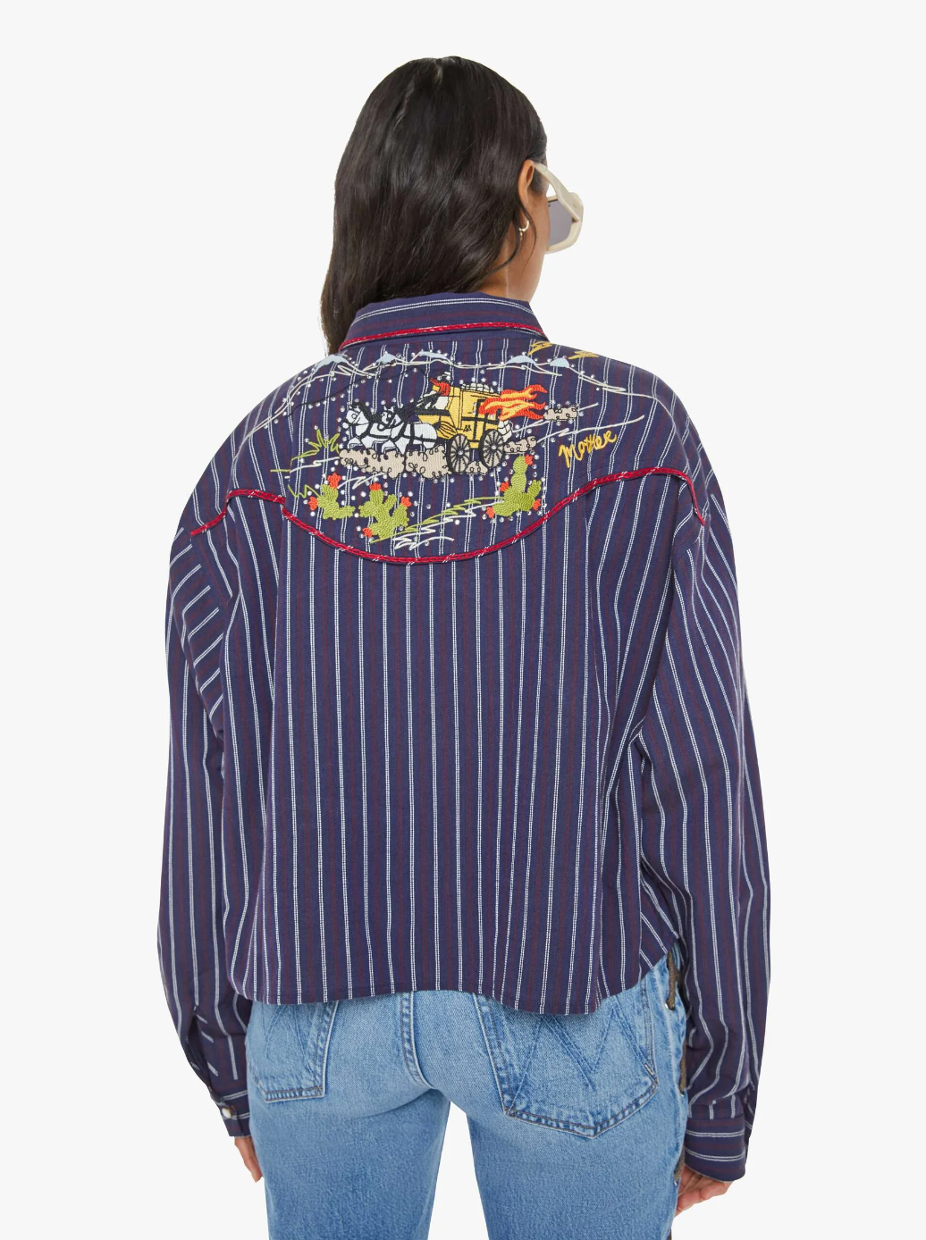 The person with long hair is seen wearing "The Riding Shotgun" by Mother—a navy cropped striped jacket paired with light blue jeans. The back of the jacket features cowboy-inspired embroidery depicting a landscape with a setting sun, as the person faces away from the camera.