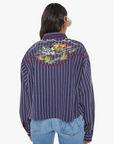 The person with long hair is seen wearing "The Riding Shotgun" by Mother—a navy cropped striped jacket paired with light blue jeans. The back of the jacket features cowboy-inspired embroidery depicting a landscape with a setting sun, as the person faces away from the camera.