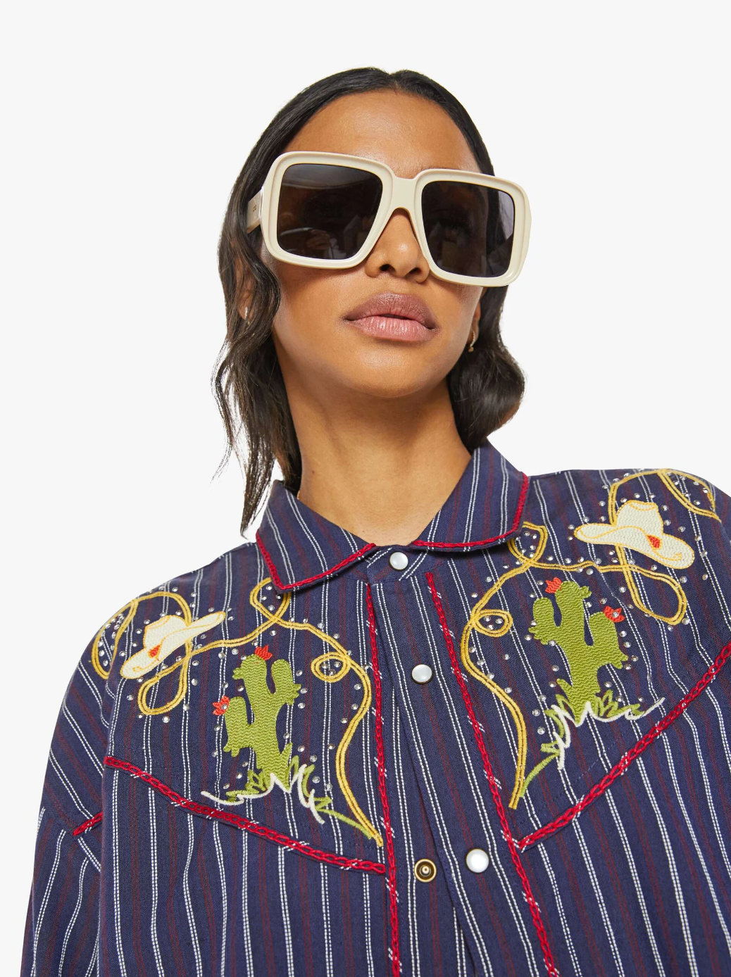 A person wearing large white sunglasses and a shirt named "The Riding Shotgun" by Mother, featuring colorful cactus embroidery and a striped Western button-up design with pearl snaps that enhance its cowboy charm against a plain white background.