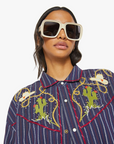 A person wearing large white sunglasses and a shirt named "The Riding Shotgun" by Mother, featuring colorful cactus embroidery and a striped Western button-up design with pearl snaps that enhance its cowboy charm against a plain white background.