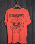 A red Ramones Crew Tee by Made Worn hangs on a wooden hanger against a dark wooden wall. Made in the USA from soft cotton, the back features "RAMONES" along with a circular emblem displaying the names "Johnny," "Joey," "Dee Dee," and "Tommy" encircling a stylized eagle.