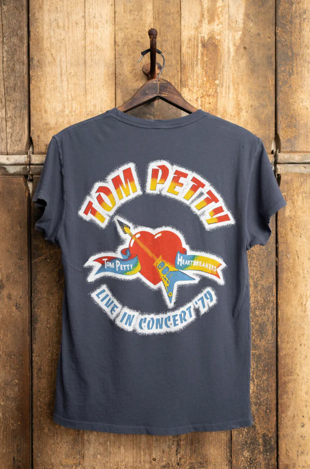 A Tom Petty 1979 Crew Tee from Made Worn is displayed on a wooden wall, featuring a vintage unisex fit and a graphic that reads "Tom Petty & the Heartbreakers," with a red heart pierced by a yellow guitar. Below, it says "Live in Concert '79," proudly made in the USA.