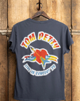 A Tom Petty 1979 Crew Tee from Made Worn is displayed on a wooden wall, featuring a vintage unisex fit and a graphic that reads "Tom Petty & the Heartbreakers," with a red heart pierced by a yellow guitar. Below, it says "Live in Concert '79," proudly made in the USA.