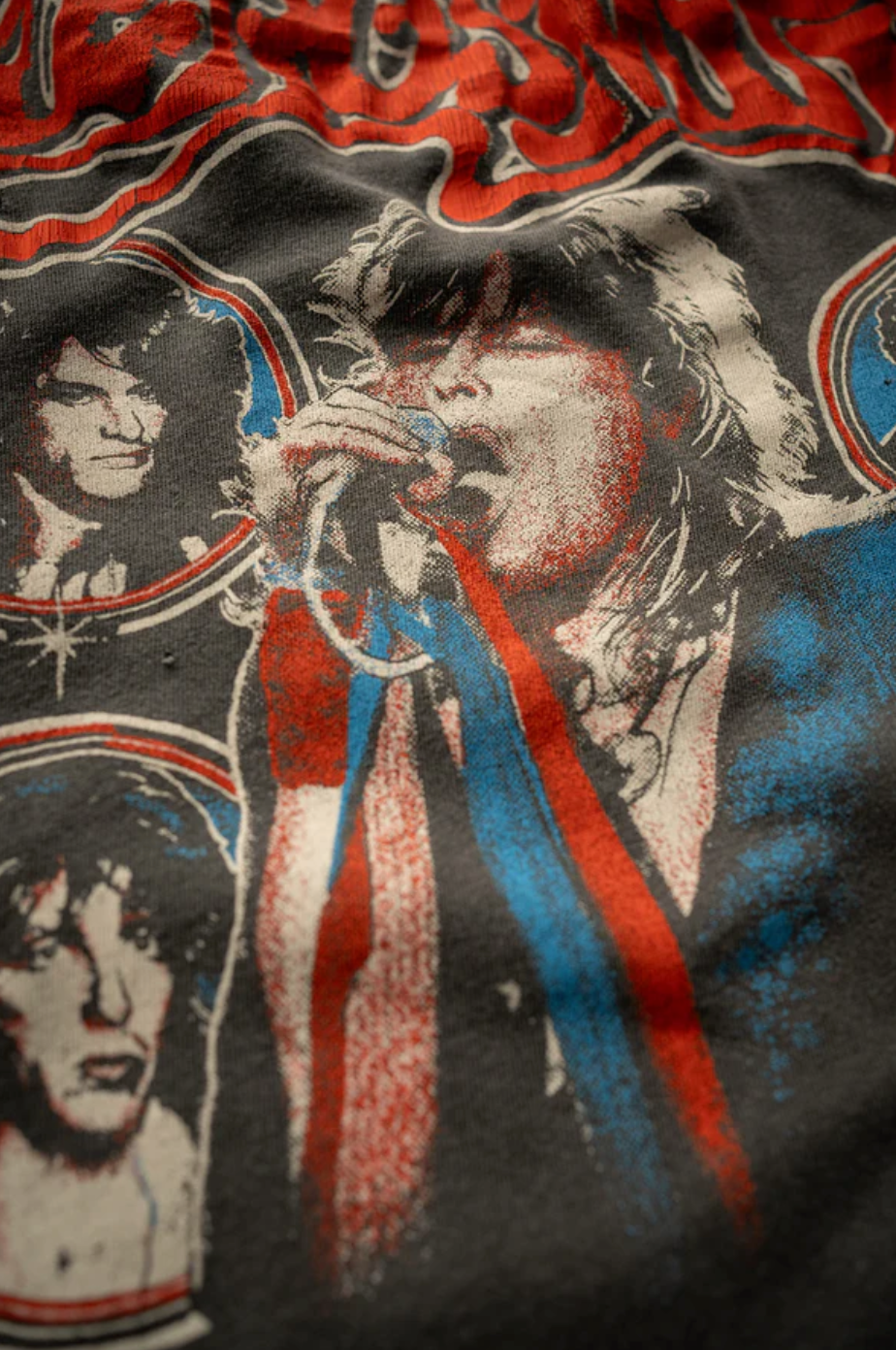 The Aerosmith In Concert Crew Tee by Made Worn is a vintage unisex fit t-shirt with a 1970s rock band design. It features unique distressing that highlights the lead singer with a microphone, surrounded by smaller portraits of other band members. The tee predominantly uses red, blue, and white colors to create an authentic retro vibe. Made in USA.