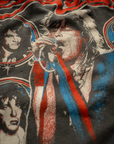 The Aerosmith In Concert Crew Tee by Made Worn is a vintage unisex fit t-shirt with a 1970s rock band design. It features unique distressing that highlights the lead singer with a microphone, surrounded by smaller portraits of other band members. The tee predominantly uses red, blue, and white colors to create an authentic retro vibe. Made in USA.