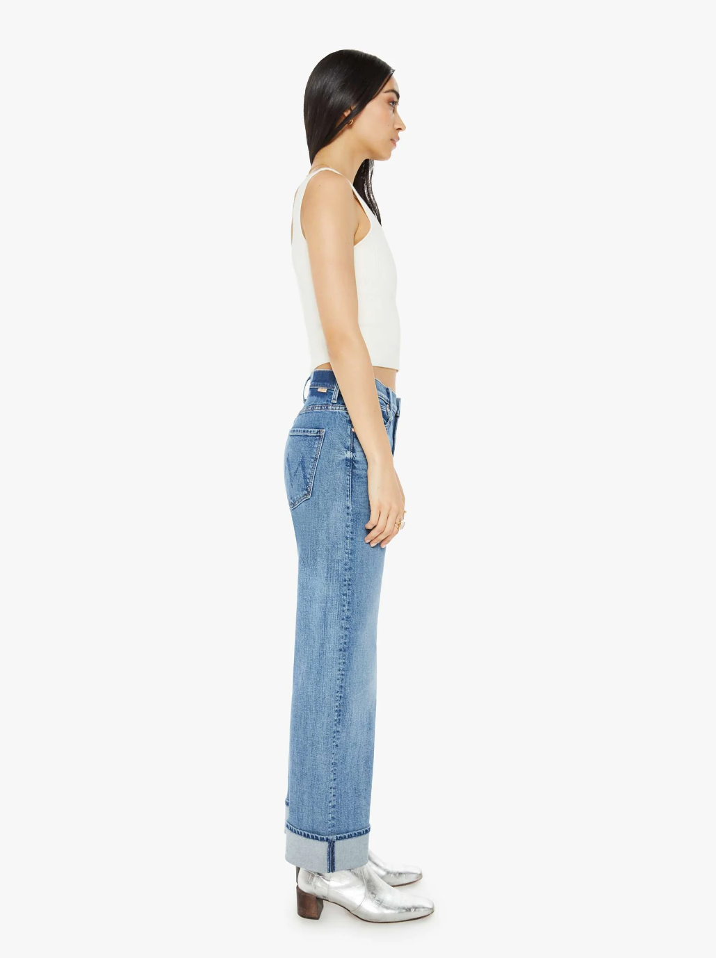 A person with long dark hair stands in profile, showcasing The Dodger Flood Cuff from Mother, a white sleeveless top, and blue high-waisted jeans with loose straight legs rolled at the cuffs. Silver ankle boots complete the ensemble against a plain white backdrop.