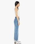 A person with long dark hair stands in profile, showcasing The Dodger Flood Cuff from Mother, a white sleeveless top, and blue high-waisted jeans with loose straight legs rolled at the cuffs. Silver ankle boots complete the ensemble against a plain white backdrop.