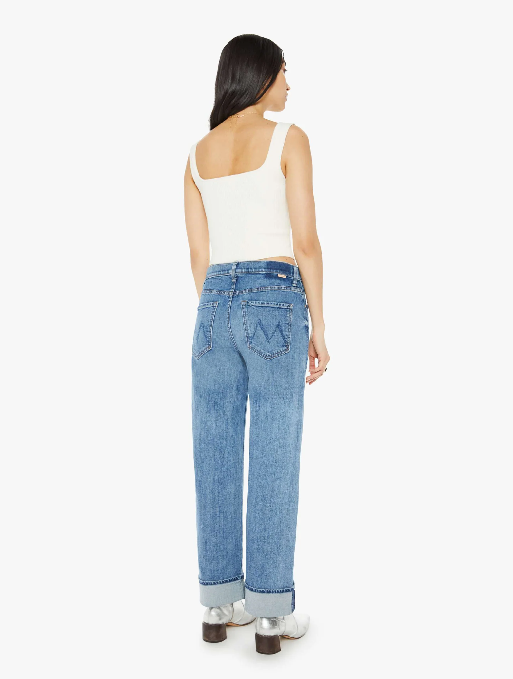 A person with long dark hair is seen from behind wearing The Dodger Flood Cuff by Mother, a pair of loose-fitting, faded light-wash denim jeans with cuffed hems, paired with a white sleeveless top and silver shoes. They are standing against a plain white background.