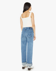 A person with long dark hair is seen from behind wearing The Dodger Flood Cuff by Mother, a pair of loose-fitting, faded light-wash denim jeans with cuffed hems, paired with a white sleeveless top and silver shoes. They are standing against a plain white background.