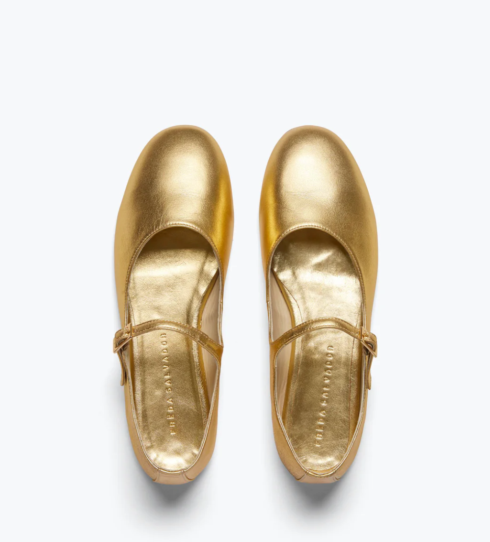 The AUBREY Flat from Freda Salvador, a pair of shiny gold ballet flats made from Italian leather and featuring a padded leather footbed, are elegantly positioned side by side on a pristine white background.