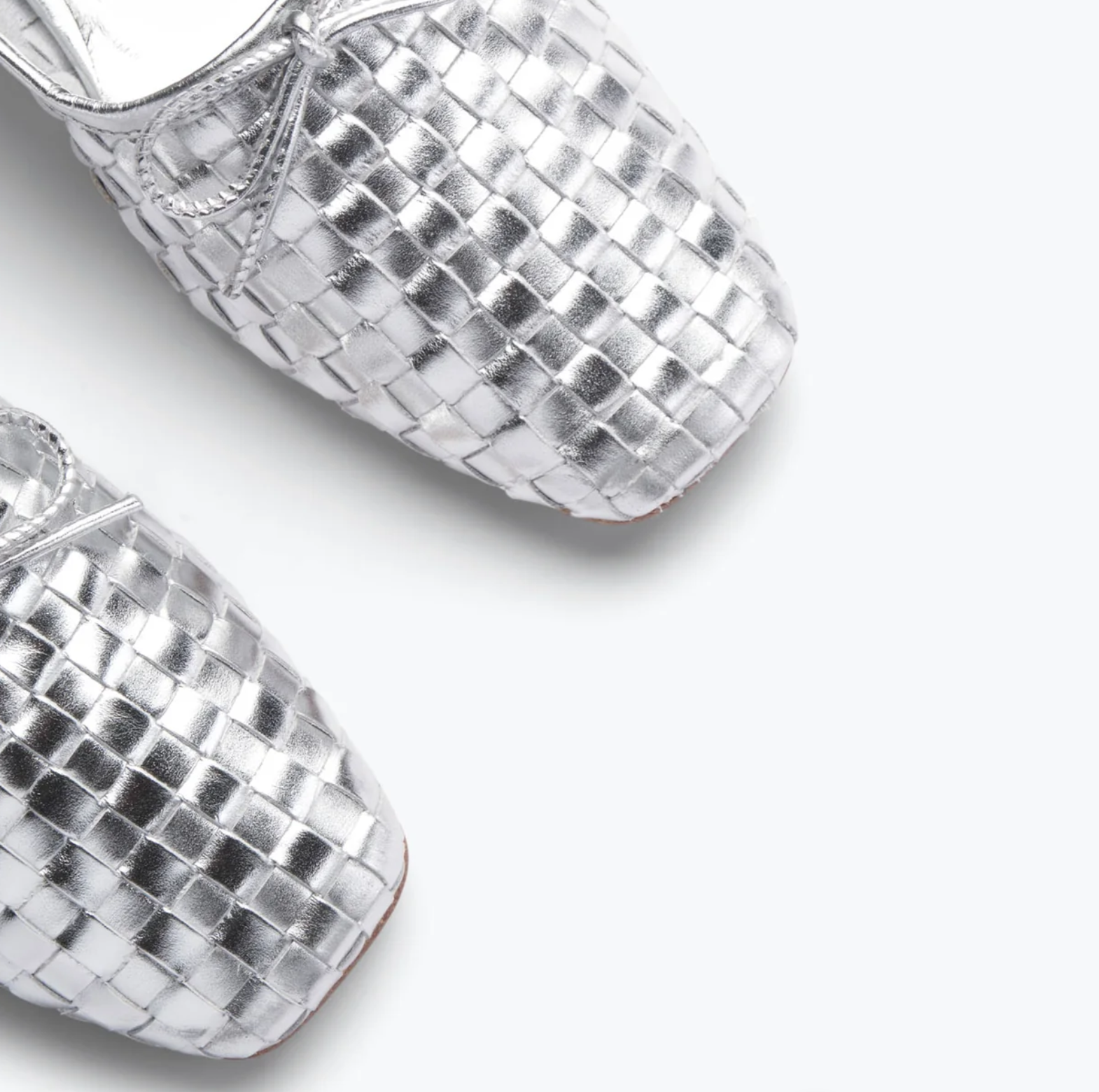 Close-up of elegant JADA Flat ballet shoes on a white background. These exquisite silver woven-texture flats, crafted by Freda Salvador, boast a shiny, metallic finish and feature a simple bow detail at the top, highlighting the luxurious quality of Spanish leather craftsmanship.