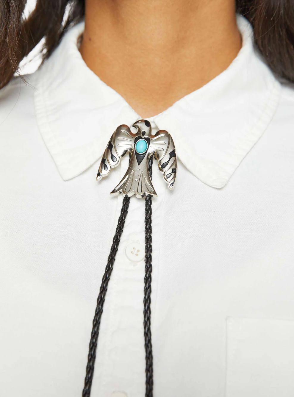 A person is wearing a white collared shirt accented by "Be On The Look Out" bolo tie from Mother, featuring a silver bird silhouette and faux turquoise stone, elegantly fastened just below the collar.