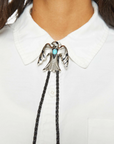 A person is wearing a white collared shirt accented by "Be On The Look Out" bolo tie from Mother, featuring a silver bird silhouette and faux turquoise stone, elegantly fastened just below the collar.