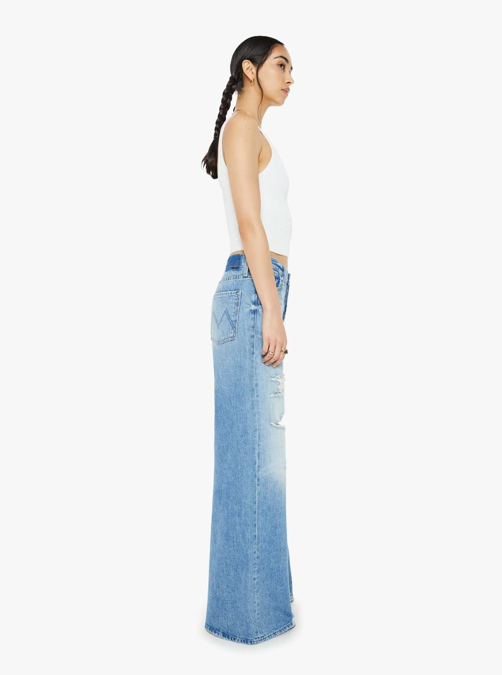 A person with braided hair is dressed in a white sleeveless top and high-rise, wide-leg distressed jeans from Mother, known as The Ditcher Roller Sneak. They are posed sideways against a plain white background.