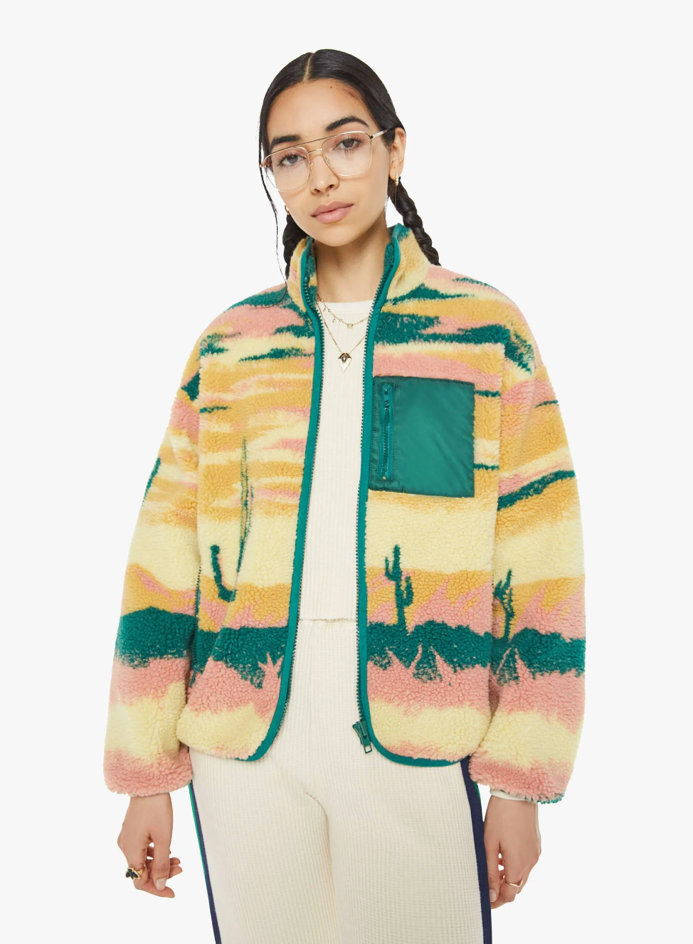 A person with glasses is wearing "The Heat Lamp Cactus Cooler" sherpa jacket by Mother, featuring a desert sunset graphic in vibrant green, pink, and yellow hues. The jacket is complemented by stylish balloon sleeves and a green chest pocket. They have long dark hair and are dressed in matching light-colored pants.