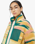 A person with long dark hair in braids is wearing glasses and The Heat Lamp Cactus Cooler, a colorful, patterned fleece jacket from Mother, featuring balloon sleeves. The jacket boasts a high collar and a teal chest pocket, artfully capturing the vibrant hues of a desert sunset graphic against the white background.