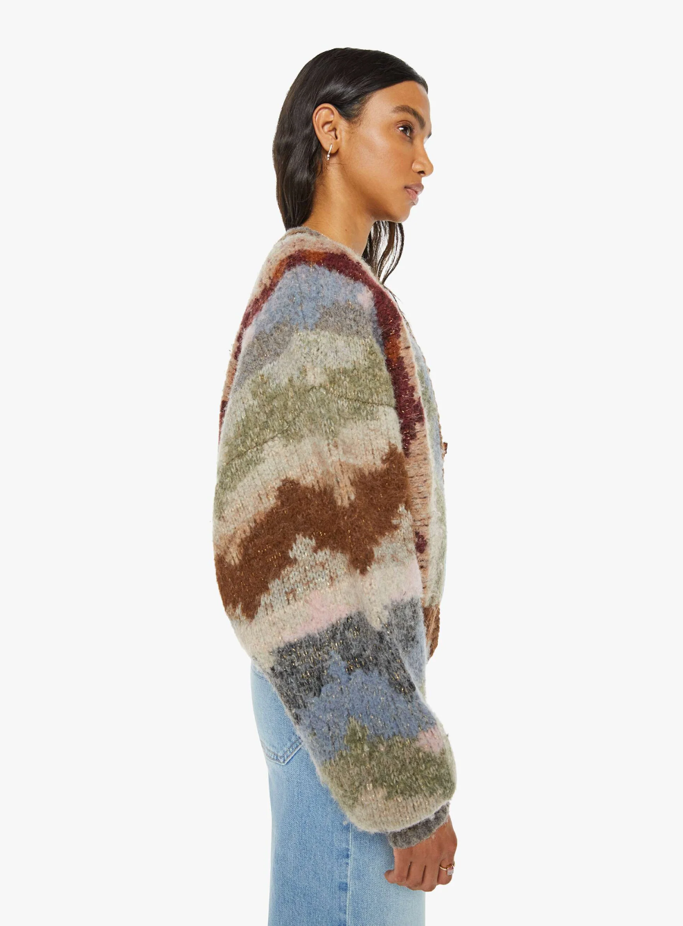 A woman stands in profile against a white background, adorned in The Soaring High Cardigan by Mother, a colorful and fuzzy wool and alpaca blend sweater featuring abstract pastel prints in brown, blue, gray, and pink shades. She pairs it with light blue jeans, her dark hair flowing freely while she accessorizes with small hoop earrings.