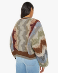 A person with long dark hair, seen from the back, is wearing The Soaring High Cardigan by Mother. This chunky sweater, made from a blend of wool and alpaca, features a fuzzy crewneck design with an abstract pattern in earthy tones of brown, green, blue, and red. It is stylishly paired with light blue jeans.